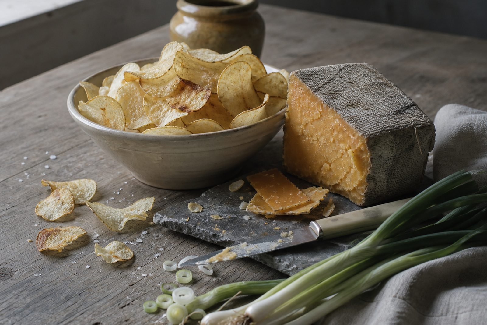 A cream stoneware bowl of pale cupped crisps on a weathered plank table, beside a cloth-bound cheddar truckle with a cut face, thin shavings of cheese and a bone-handled knife on a slate board, sliced spring onions, a stoneware jar behind and broken crisps and salt at the left.