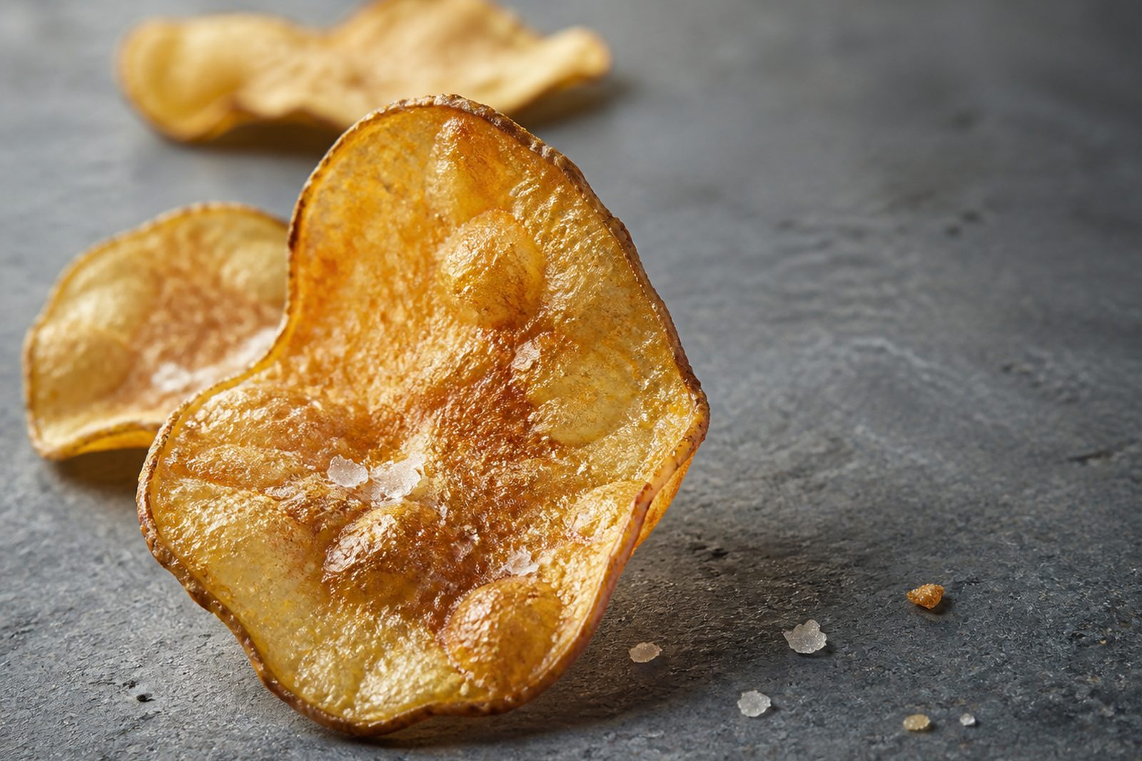 Extreme close-up of a single golden hand-cooked crisp standing on grey slate, its surface covered in irregular blistered domes and a rippled darker-flecked rim, with coarse salt crystals embedded in the oil and two softly out-of-focus crisps behind it.