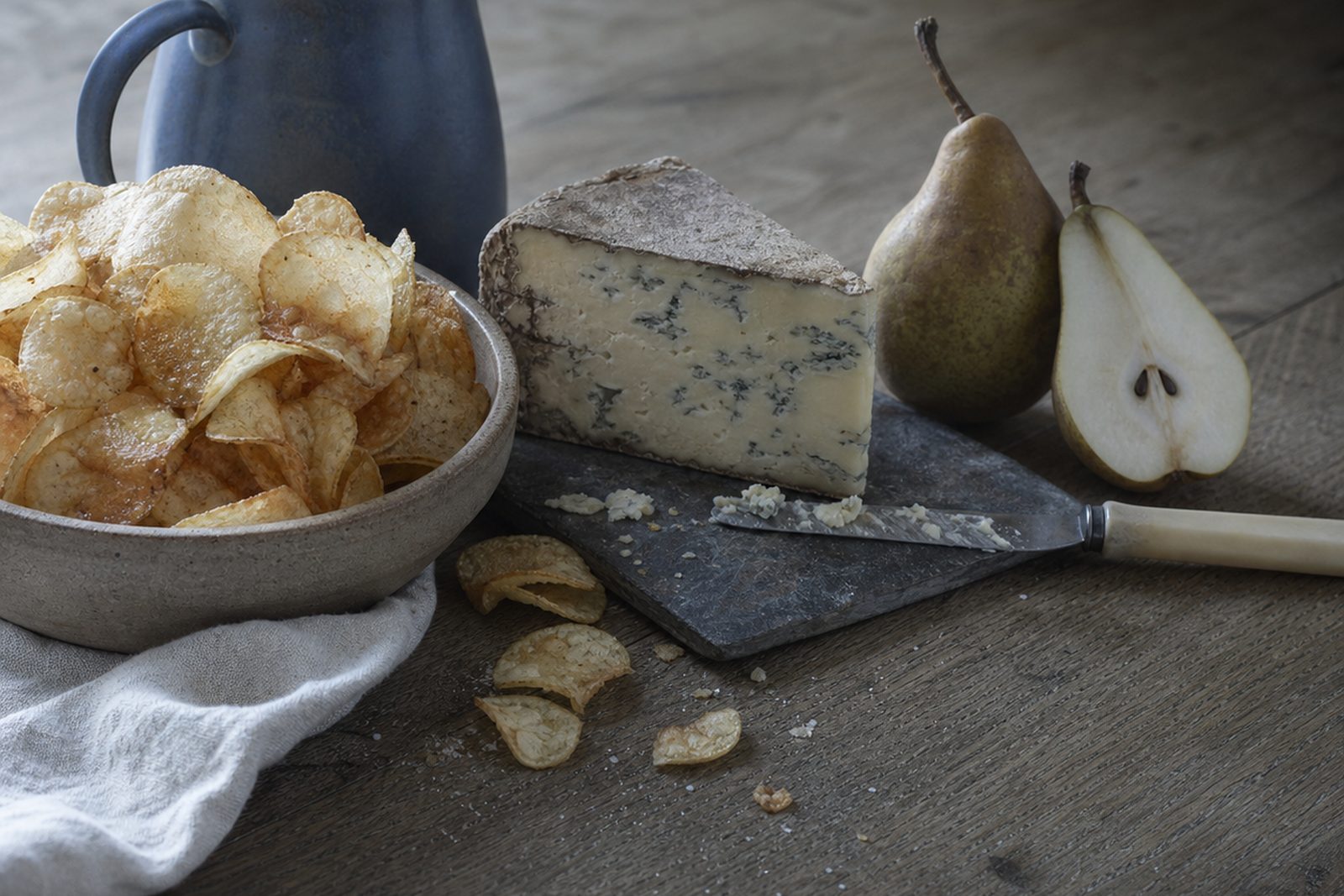 A stone bowl of thin cupped crisps beside a blue stoneware jug on a grey wooden table, with a crusted wedge of blue-veined Stilton on a slate board, a bone-handled knife carrying crumbled cheese, a whole pear and a halved pear, and fallen crisps on the boards below.