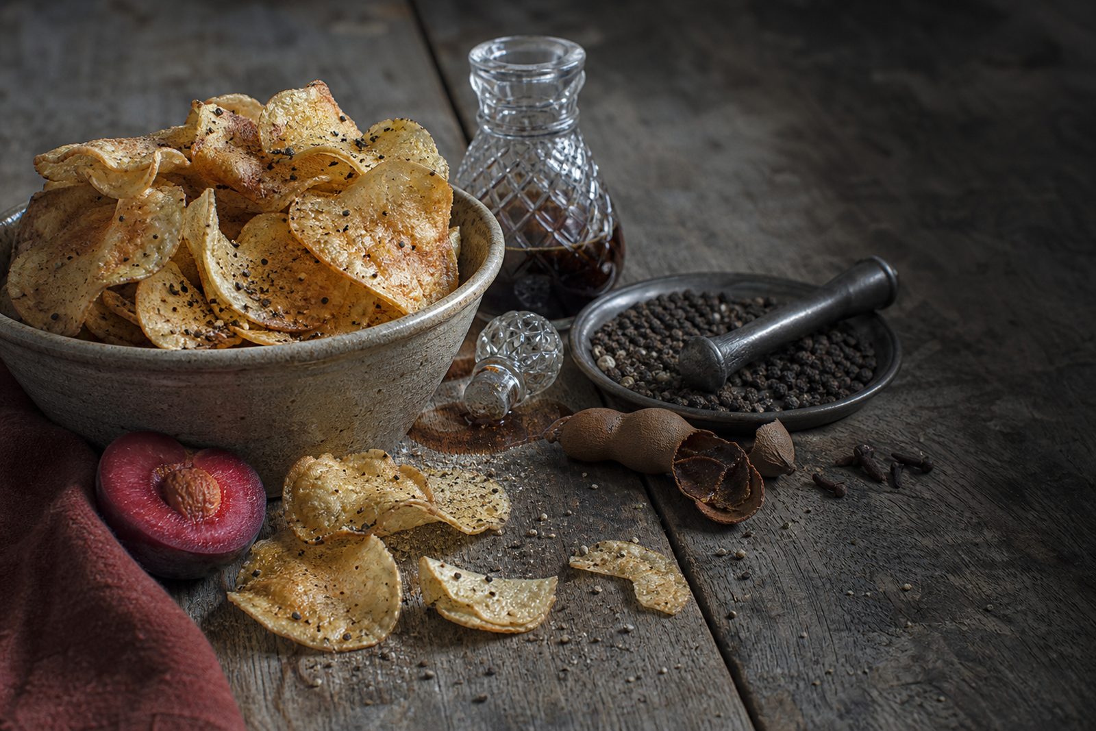 A stone bowl of crisps dusted with cracked black pepper on dark weathered boards, with a cut-crystal decanter of dark sauce, a pewter dish of peppercorns and pestle, tamarind pods, cloves, a halved plum on a dark red cloth and fallen peppered crisps in the foreground.