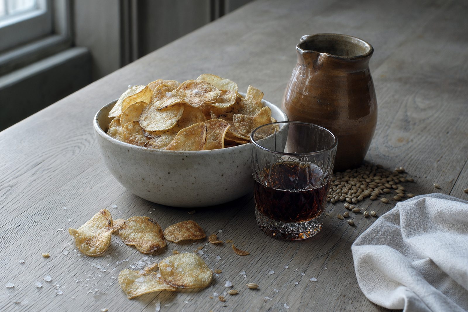 A speckled stone bowl piled with thin salted crisps on a pale scrubbed table beside a cold window, with a brown salt-glazed jug, a cut-crystal tumbler of dark malt vinegar, loose barley grains, a linen cloth and fallen crisps and salt scattered across the wood.